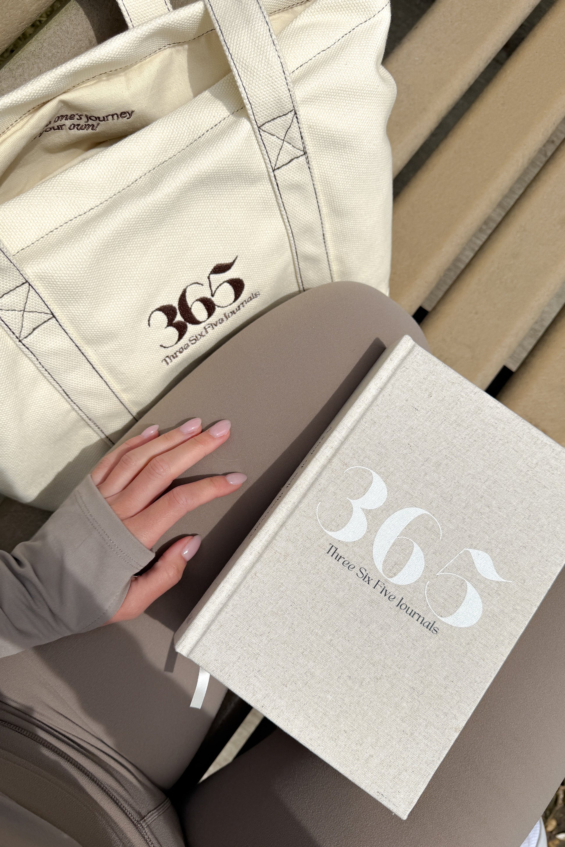 Journal with '365' branding held by a hand, next to a beige tote bag on a wooden surface.
