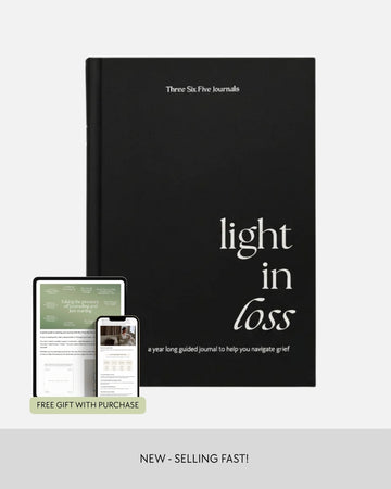 A black journal titled 'light in loss' by Three Six Five Journals, shown upright with a tablet and smartphone displaying the included guided journal content. The image includes a 'FREE GIFT WITH PURCHASE' banner and 'NEW - SELLING FAST!' text at the bottom.