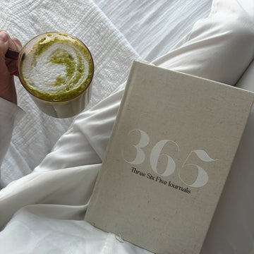 person holding a matcha coffee and the 365 Journal on their lap on a white bed 