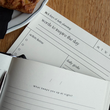 An open lined journal on a wooden table with a pastry on a plate at the top left. A second sheet of paper with writing prompts is partially visible underneath.