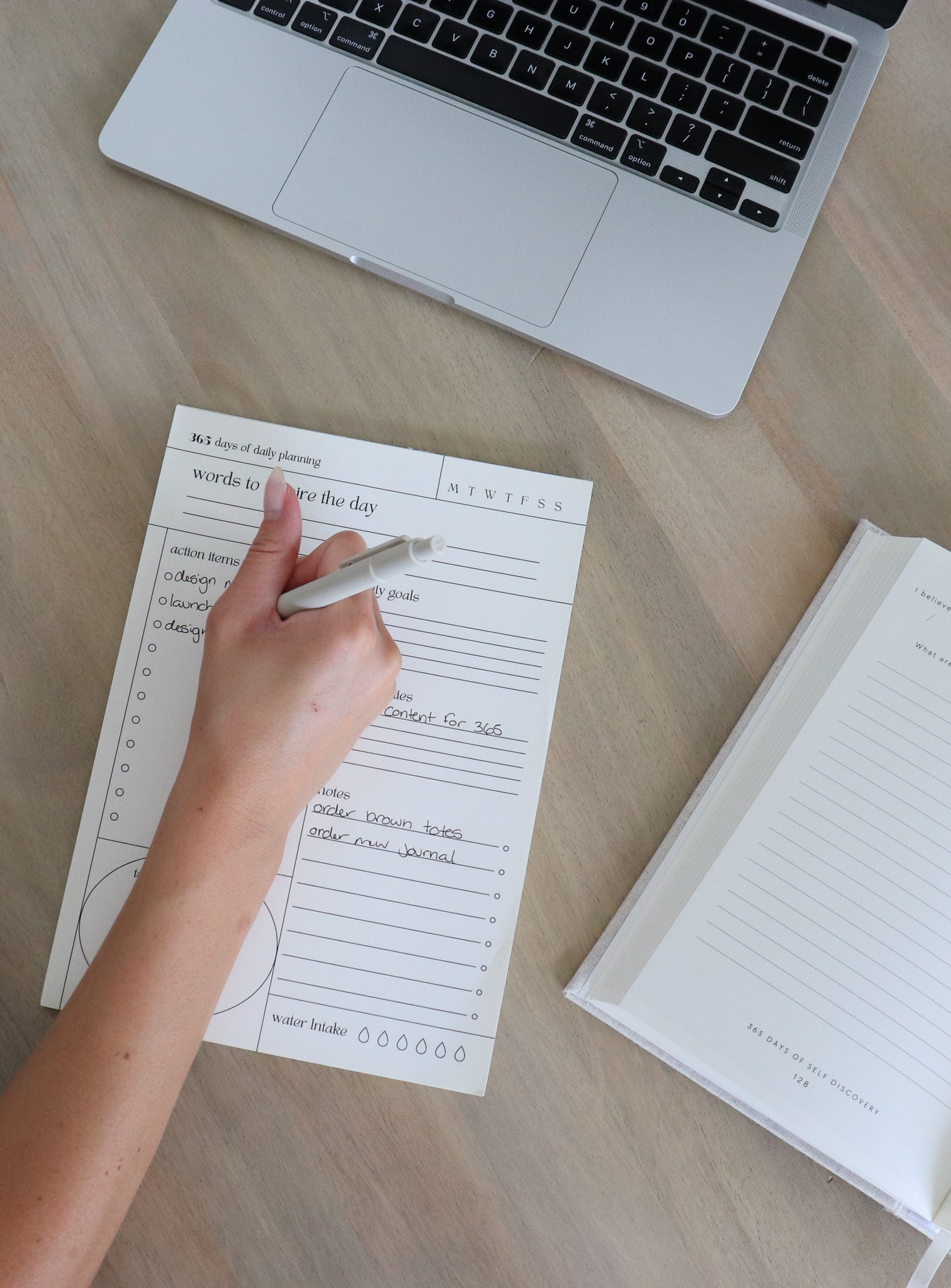 Person writing on the Daily Align notepad with a laptop and another planner on a wooden surface